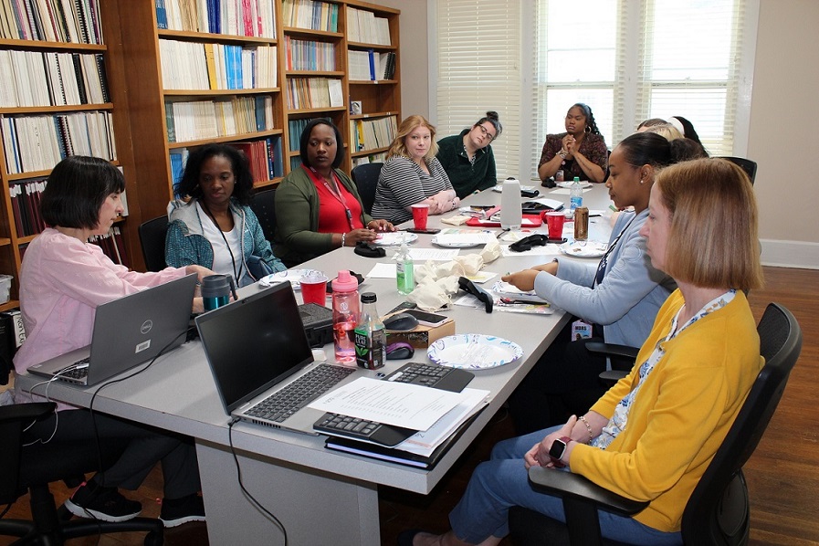 A group of professionals seated around a conference table in a bright room with notebooks, laptops, and drinks on the table. 