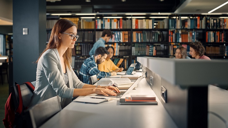 Image of a woman wearing glasses at a library typing on her laptop.