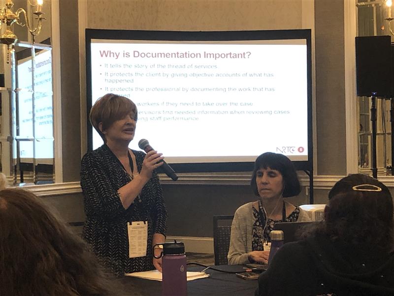 Jennifer standing and speaking into a microphone during the "Did I Really Do It?:The Importance of Documentation" presentation. Kendra is sitting beside her.