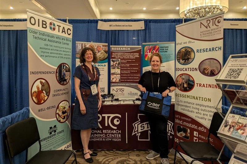 NRTC booth at the AER Conference. Polly Abbott and Jamie Boydstun, staff members at the NRTC, are standing beside the booth. Polly is wearing a long blue dress and Jamie is holding an AER tote bag. Banners about OIB-TAC and the NRTC surround the booth.