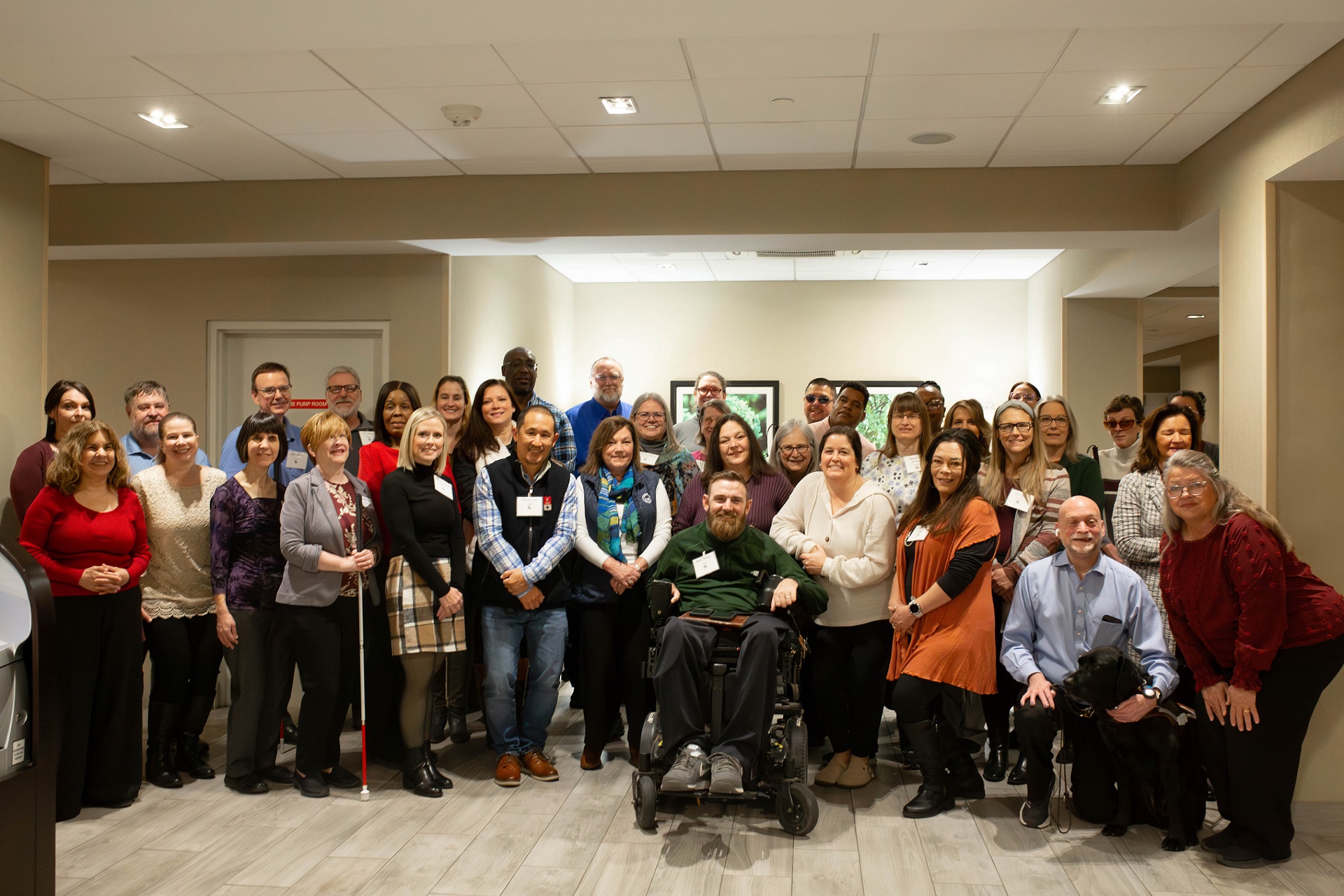 A large group of conference attendees poses for a group photo, smiling. Some individuals are using mobility aids, including a white cane and a guide dog.